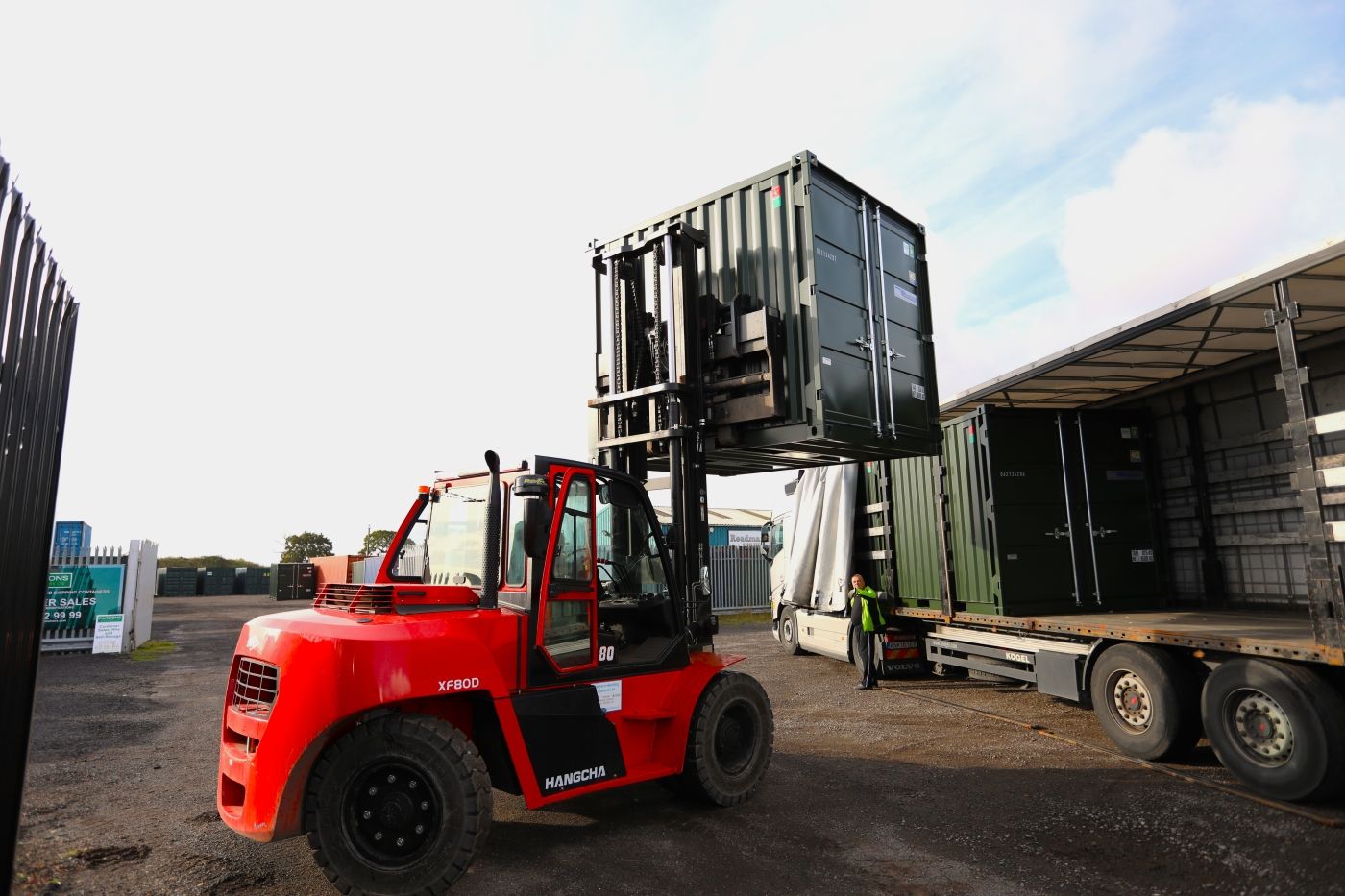A forklift loading a green shipping container onto a curtain-sided trailer